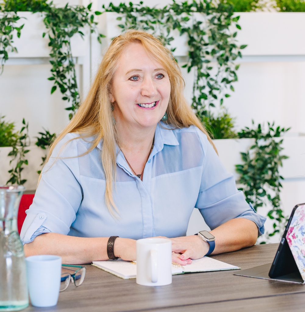Image shows me, a lady with long blonde hair wearing a pale blue shirt sitting at a desk with a laptop and notebook.